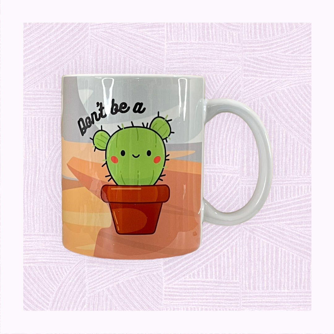 Ceramic mug with printed desert background, a cute cactus character with the phrase ‘Don’t be a…’ above it.