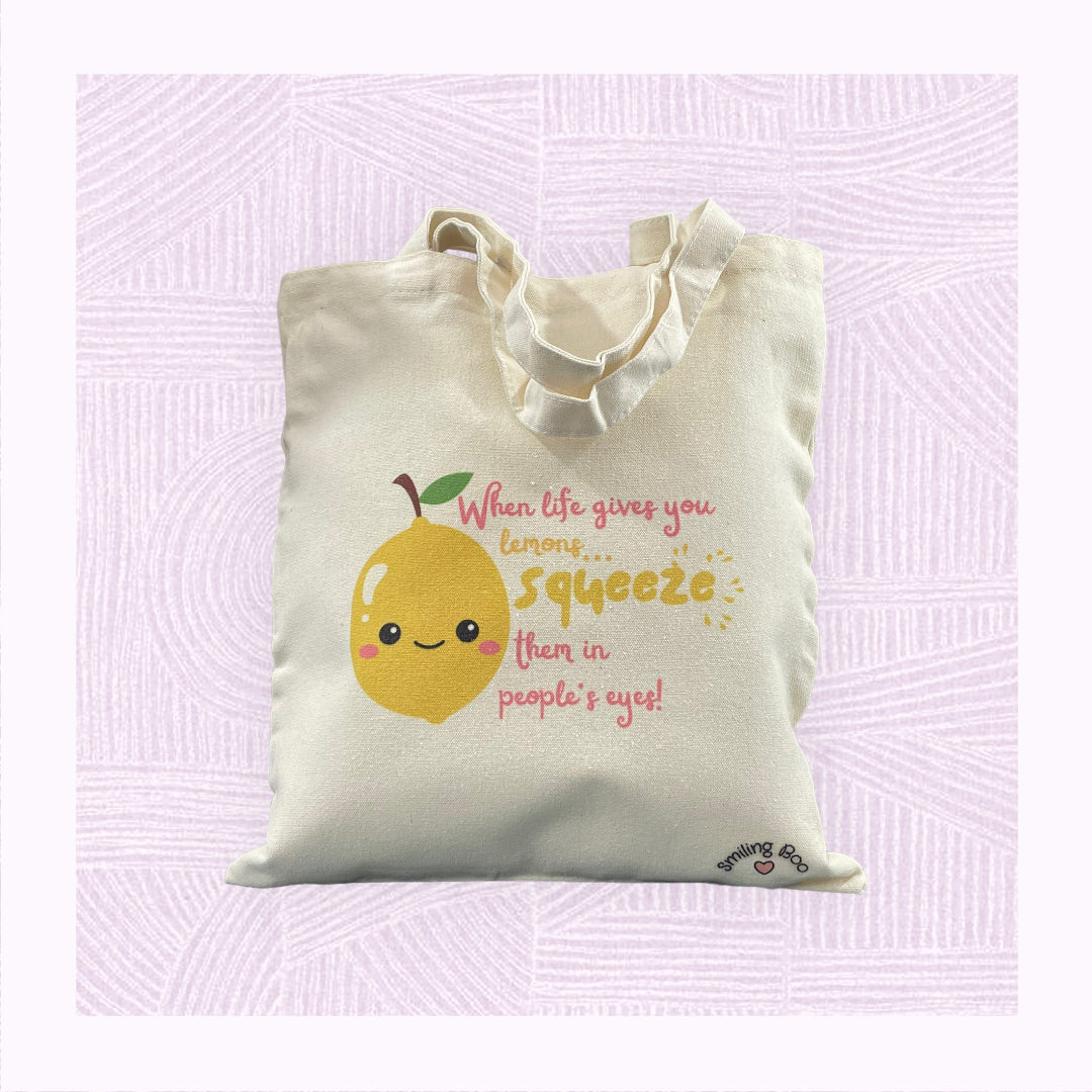 Canvas tote bag with a cute lemon character in the foreground with the phrase ‘When life gives you lemons, squeeze them in people’s eyes’.