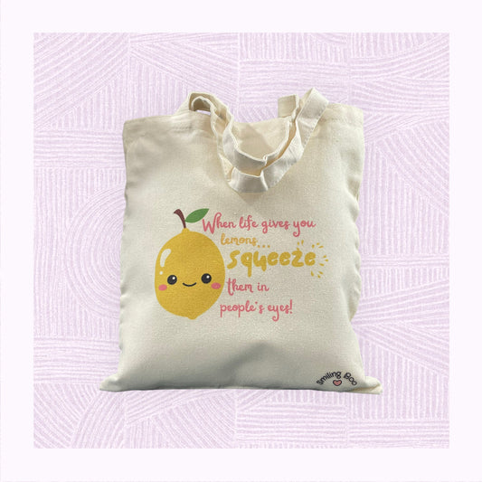Canvas tote bag with a cute lemon character in the foreground with the phrase ‘When life gives you lemons, squeeze them in people’s eyes’.